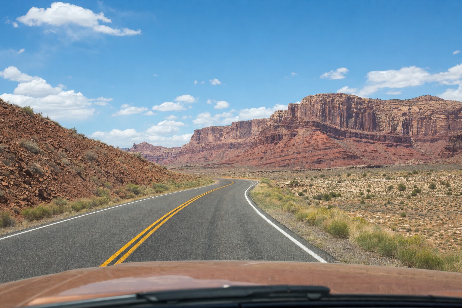 A driver's view of a paved scenic road curving toward layered red rock cliffs
